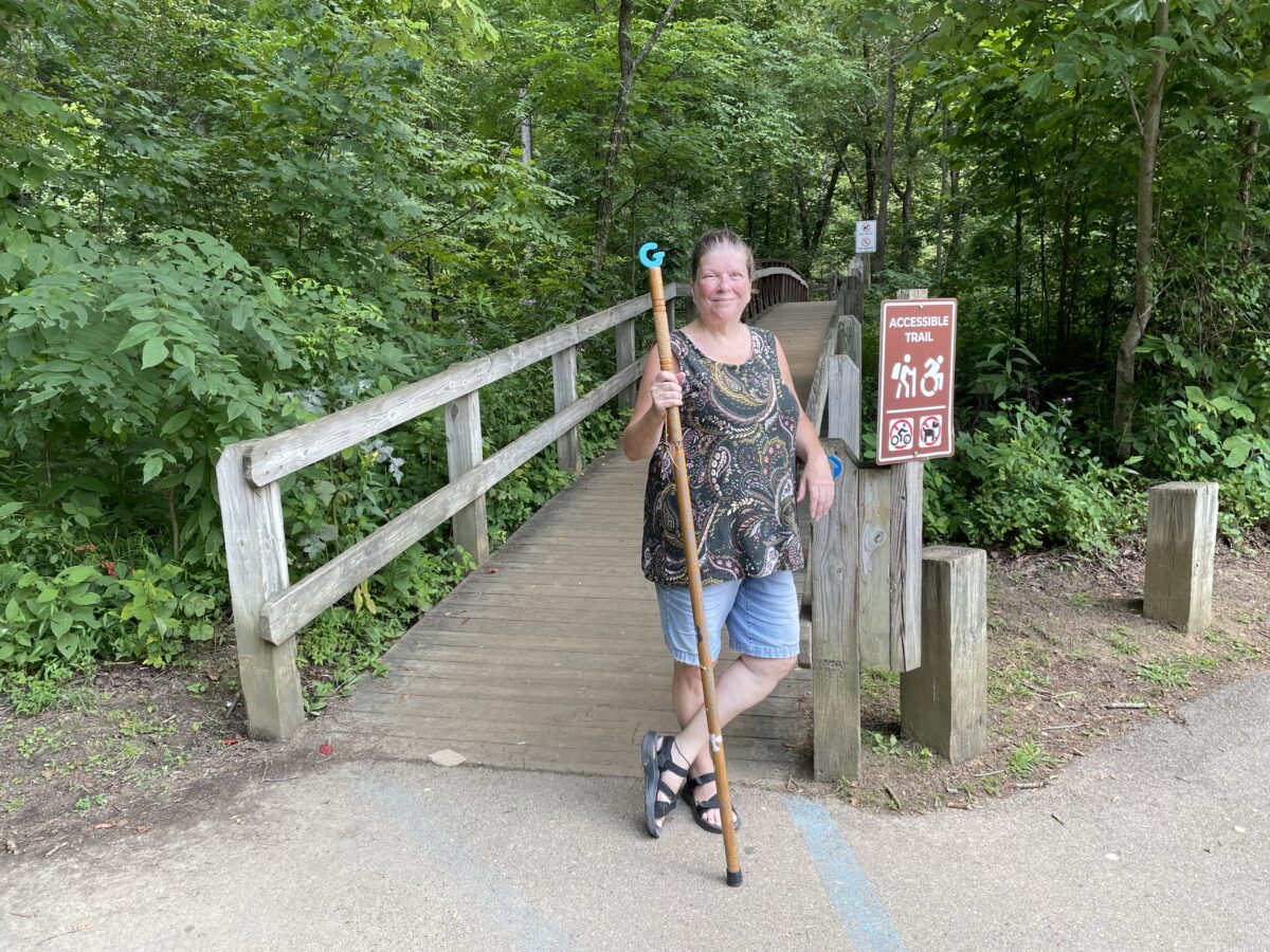 Grammy standing with her walking stick next to a wooden bridge. A sign next to her says "Accessible Trail" and shows icons of a hiker and the modern Wheelchair Symbol.